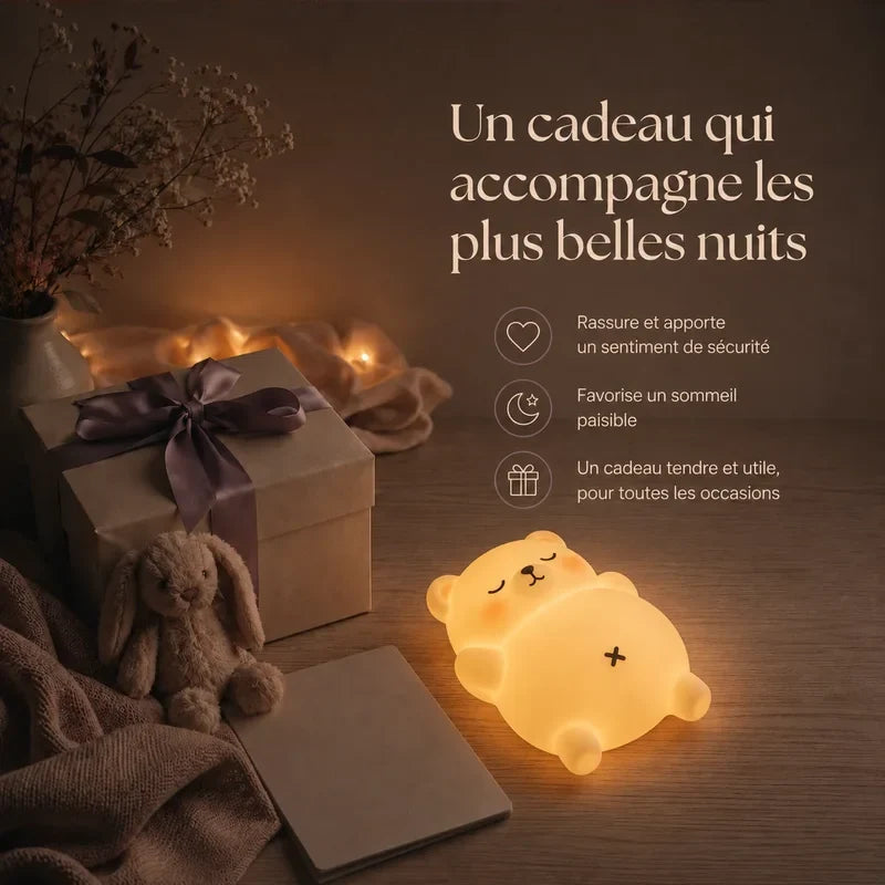 Veilleuse en forme d’ours allongé sur le dos présentée dans une ambiance cadeau avec une boîte et un ruban.
