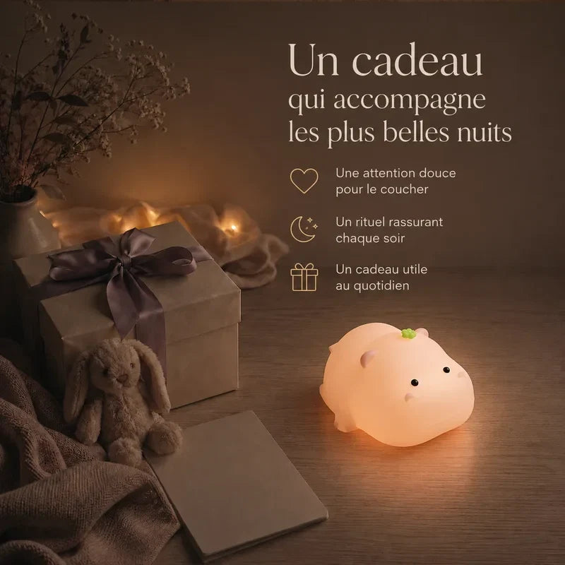 Veilleuse rose en forme d’hippopotame rond présentée dans une ambiance cadeau avec une boîte et un ruban.