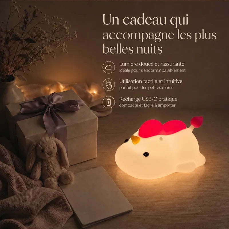 Veilleuse en forme de licorne allongée à crinière rose présentée dans une ambiance cadeau avec une boîte et un ruban.