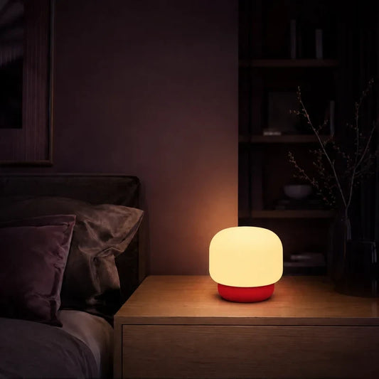 Lampe arrondie avec base rouge allumée sur une table de chevet dans une ambiance nocturne.
