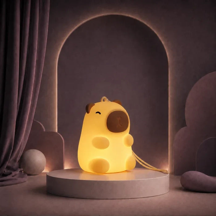 Veilleuse nomade en forme de capybara avec dragonne allumée sur un podium mauve.