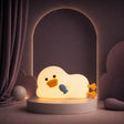 Veilleuse en forme de canard allongé avec un petit poisson bleu allumée sur un podium mauve.